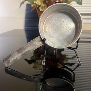 May include: A silver metal saucepan with a long handle, sitting on a black metal stand. The saucepan is empty and has a shiny surface.