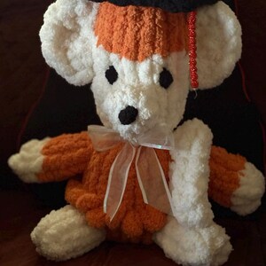 May include: A handmade plush bear in white, orange, and black, wearing a graduation cap and a sheer ribbon bow. The bear has black eyes and nose, and the cap has a red tassel. The bear is sitting on a brown surface.