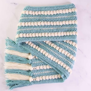 Puede incluir: Una manta de punto de crochet con un patrón a rayas en color turquesa y blanco. La manta tiene un borde de flecos y un punto de textura de pompón.