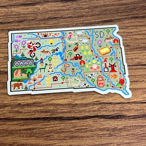 May include: A pixel art style sticker of the state of South Dakota with the abbreviation "SD" in the upper right corner. The sticker features various icons representing the state's culture and landscape, including a bison, a tractor, a fish, a cornfield, and a prairie dog.