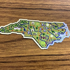 Puede incluir: Una pegatina con un mapa pixelado de Carolina del Norte con detalles verdes, azules y rojos. El mapa incluye iconos que representan puntos de referencia y las letras "NC". La pegatina está sobre una superficie de madera.