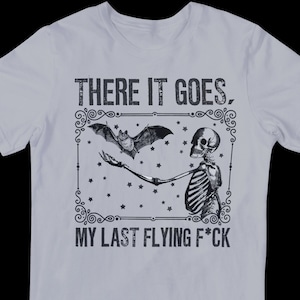 May include: A light gray t-shirt with a black and white graphic of a skeleton holding a bat. The graphic is surrounded by a decorative frame and stars. The text "THERE IT GOES. MY LAST FLYING F*CK" is printed below the graphic.