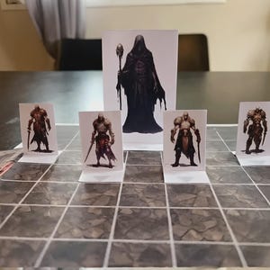 Puede incluir: Un conjunto de cinco miniaturas de papel para juegos de mesa con personajes de fantas&iacute;a, incluyendo cuatro guerreros con armadura y una figura central encapuchada con un bast&oacute;n, sobre un mapa de cuadr&iacute;cula con patr&oacute;n de piedra.