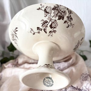 Pode incluir: Uma tigela de pedestal de cer&acirc;mica branca com um design floral castanho. A tigela tem uma base larga e rasa e um caule alto e fino. A parte inferior da tigela tem um carimbo castanho com o texto "MADE IN ENGLAND,  BOULTONS,  LIMITED,  BURSLEM".