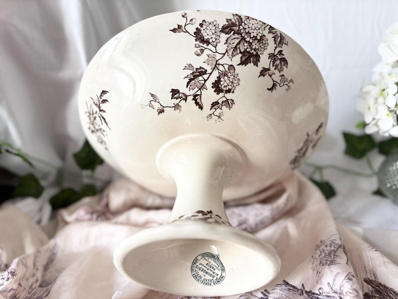 Pode incluir: Uma tigela de pedestal de cer&acirc;mica branca com um design floral castanho. A tigela tem uma base larga e rasa e um caule alto e fino. A parte inferior da tigela tem um carimbo castanho com o texto "MADE IN ENGLAND,  BOULTONS,  LIMITED,  BURSLEM".