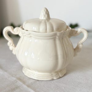 Op de afbeelding: Crèmekleurige keramische suikerpot met deksel. De kom heeft een afgeronde vorm met verticale ribbels en sierlijke handvatten. Het deksel is bekroond met een decoratieve knop. De suikerpot staat op een wit oppervlak.