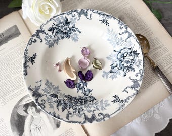 May include: A round ceramic plate with a cream-colored base and a dark blue floral pattern featuring intricate botanical illustrations and scrollwork. The plate contains several small dried flower petals in shades of purple and tan.