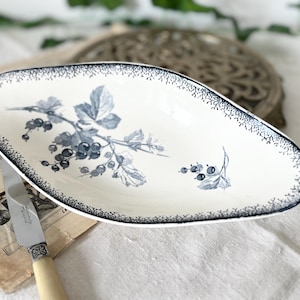 Może przedstawiać: Owalny biały ceramiczny półmisek z czarnym wzorem jagód i liści. Obrzeże zdobione czarnym wzorem w kropki. Obok leży zabytkowy nóż z kremową rączką.