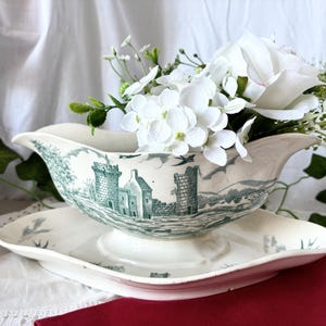 Könnte beinhalten: Eine weiße Porzellan-Sauciere mit einem grün-weißen Schlossdesign. Die Sauciere ist mit weißen Blumen gefüllt und steht auf einer passenden Untertasse.