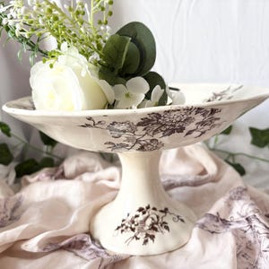 Pode incluir: Uma tigela de cer&acirc;mica branca com pedestal e um padr&atilde;o floral castanho. A tigela &eacute; decorada com um delicado desenho floral e &eacute; perfeita para exibir flores ou outros itens decorativos.
