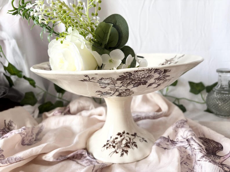 Pode incluir: Uma tigela de cer&acirc;mica branca com pedestal e um padr&atilde;o floral castanho. A tigela &eacute; decorada com um delicado desenho floral e &eacute; perfeita para exibir flores ou outros itens decorativos.