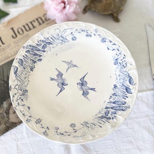 Puede incluir: Un plato de cerámica blanca con un diseño floral y de pájaros azules. El plato tiene un borde festoneado y está decorado con pájaros volando y elementos botánicos. Parte de una colección de vajilla vintage.