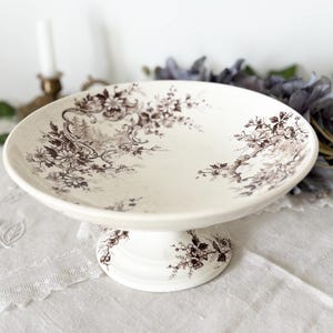 Puede incluir: Un cuenco de cerámica de color crema sobre un pedestal con un patrón de transferencia marrón oscuro de intrincados motivos florales y follaje en espiral. El cuenco tiene una base ancha y poco profunda y una base circular resistente.