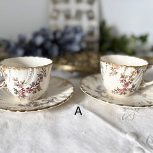 以下が含まれることがあります： 2つのアンティークティーカップとソーサー。カップとソーサーはクリーム色で、ピンクとゴールドの花柄模様です。カップとソーサーの縁はスカラップで、金色のトリムが施されています。カップには取っ手が付いています。