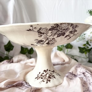 Pode incluir: Uma tigela branca com pedestal e um design floral castanho. A tigela est&aacute; sobre um fundo de tecido branco.