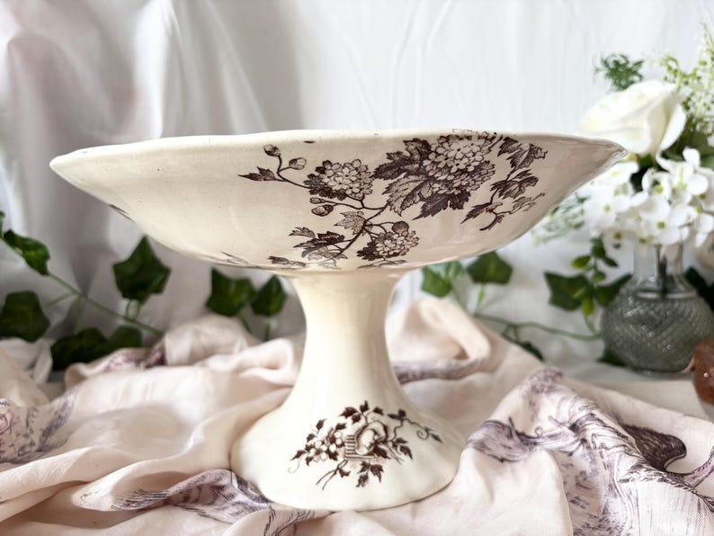 Pode incluir: Uma tigela branca com pedestal e um design floral castanho. A tigela est&aacute; sobre um fundo de tecido branco.