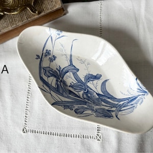 May include: An oval-shaped ceramic dish with an off-white base color and a blue botanical illustration of iris flowers and tall grasses. The letter A is printed in black to the left of the dish, which features a smooth, curved rim.
