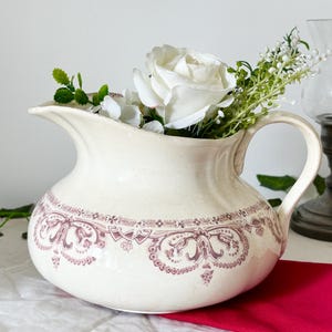Pode incluir: Uma jarra de cerâmica na cor creme com um padrão decorativo de transferência bordô ao redor da seção central. A jarra possui um corpo largo e arredondado, uma alça curva e um bico, preenchida com rosas artificiais brancas e folhagens.