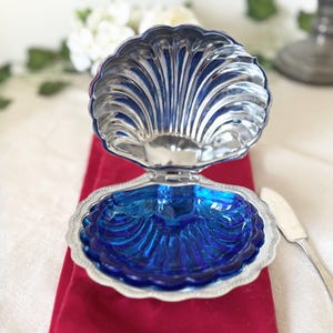 Könnte beinhalten: Eine versilberte Muschelschale mit einem Klappdeckel, der einen tiefblauen Glaseinsatz freigibt. Die Außenseite der Muschel hat eine metallische Oberfläche mit gerippten Details, während der innere Boden aus leuchtend blauem Glas besteht.