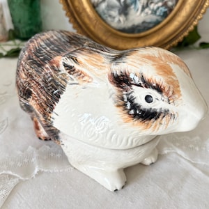 Op de afbeelding: Een keramieken cavia-figuur met een glanzende afwerking, voorzien van een driekleurige vacht met witte, tan en zwarte vlekken. Het beeldje is vormgegeven in een rustende houding met gedetailleerde vachttextuur en geschilderde ogen.