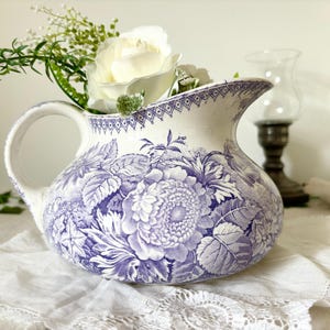 Puede incluir: Una jarra de cerámica blanca con un detallado patrón floral de color morado. El recipiente tiene un cuerpo redondeado, un asa curva y un borde geométrico decorativo en la parte superior. Está lleno de rosas blancas artificiales y follaje.