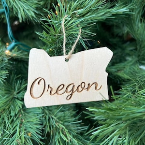 May include: A wooden ornament shaped like the state of Oregon with the word "Oregon" engraved on it. The ornament is hanging on a Christmas tree.