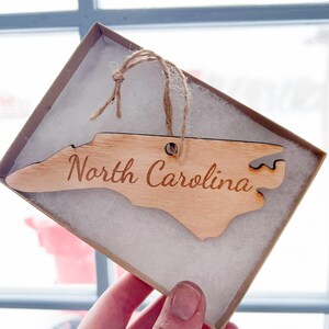 May include: A wooden ornament shaped like the state of North Carolina with the words "North Carolina" engraved on it. The ornament is in a small box with white tissue paper.