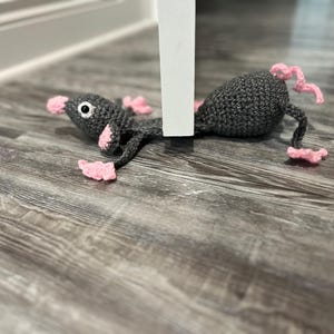 May include: A grey crocheted rat toy with pink ears and feet. The rat is lying on a wooden floor with a white doorstop behind it.