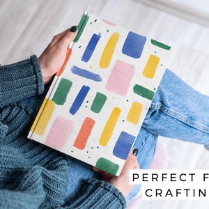 May include: A notebook with a white cover featuring colorful brushstroke designs in blue, yellow, green, pink, and orange. The text "PERFECT FOR CRAFTING" is visible on the bottom right.