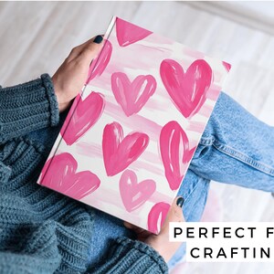 Puede incluir: Un cuaderno blanco con un patr&oacute;n de corazones rosas en acuarela. El cuaderno est&aacute; siendo sostenido por una persona que lleva un su&eacute;ter azul y jeans.
