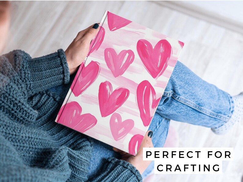 Puede incluir: Un cuaderno blanco con un patr&oacute;n de corazones rosas en acuarela. El cuaderno est&aacute; siendo sostenido por una persona que lleva un su&eacute;ter azul y jeans.