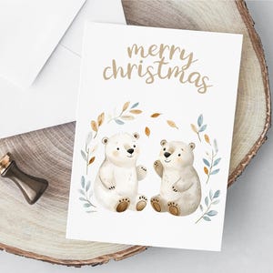 以下が含まれることがあります： 白いカードに、茶色の文字で「merry christmas」と書かれています。茶色の鼻をした白いホッキョクグマが2匹、茶色と青の葉の輪の中に並んで座っています。