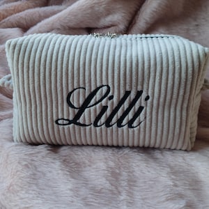Könnte beinhalten: Eine hellbeige Kosmetiktasche aus Cord mit dem Namen "Lilli" in schwarzer Schrift gestickt. Die rechteckige Tasche hat einen Reißverschluss und kleine Stoffschlaufen an jeder Seite. Die Tasche liegt auf einer rosa, flauschigen Oberfläche.