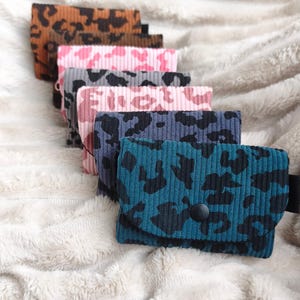 Könnte beinhalten: Eine Sammlung von fünf kleinen rechteckigen Taschen aus geripptem Cordstoff, die jeweils ein markantes Leopardenmuster in den Farben Petrol, Dunkelgrau, Hellrosa, Pink und Braun aufweisen und mit einem schwarzen Druckknopf verschlossen sind.