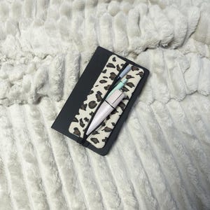 Könnte beinhalten: Ein schwarzes Notizbuch mit einem Stifthalter im Leopardenmuster und einem weißen Stift, einem hellblauen Stift und einem grauen Stift. Das Notizbuch liegt auf einer weißen, strukturierten Oberfläche.