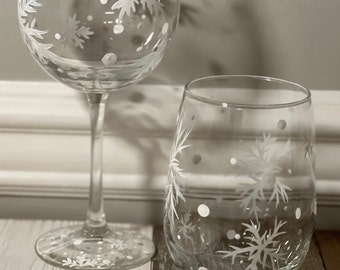 May include: Two clear wine glasses with white snowflake and dot designs. One is a stemmed wine glass, and the other is a stemless tumbler. The glasses are on a white and brown wooden surface, suggesting a winter or holiday theme.