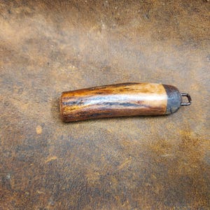 May include: A handcrafted, wooden bottle stopper with a natural, warm-toned finish. The stopper features a dark metal cap and a small metal loop for easy removal. The wood grain is visible, adding to its rustic charm.