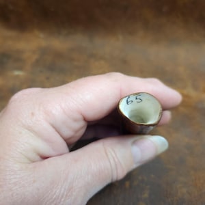 May include: A small, brown, cylindrical object with a cream-colored interior. The number "65" is written in black ink on the top rim. The object is held in a hand.