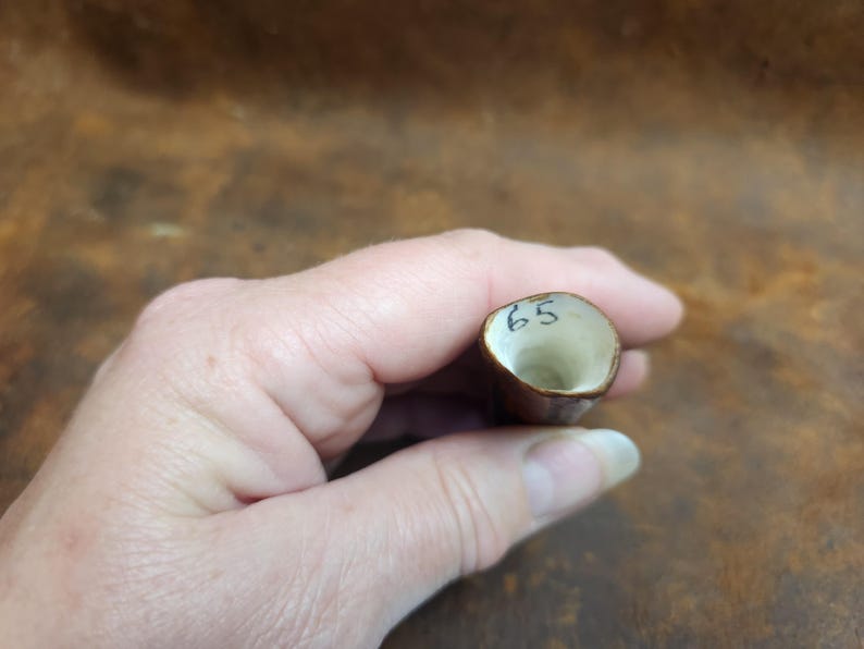 May include: A small, brown, cylindrical object with a cream-colored interior. The number "65" is written in black ink on the top rim. The object is held in a hand.