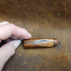 May include: A small, handcrafted wooden object with a dark brown and tan grain pattern. It has a rounded shape with a metal loop at one end. The object is held by a hand, suggesting its small size and portability.