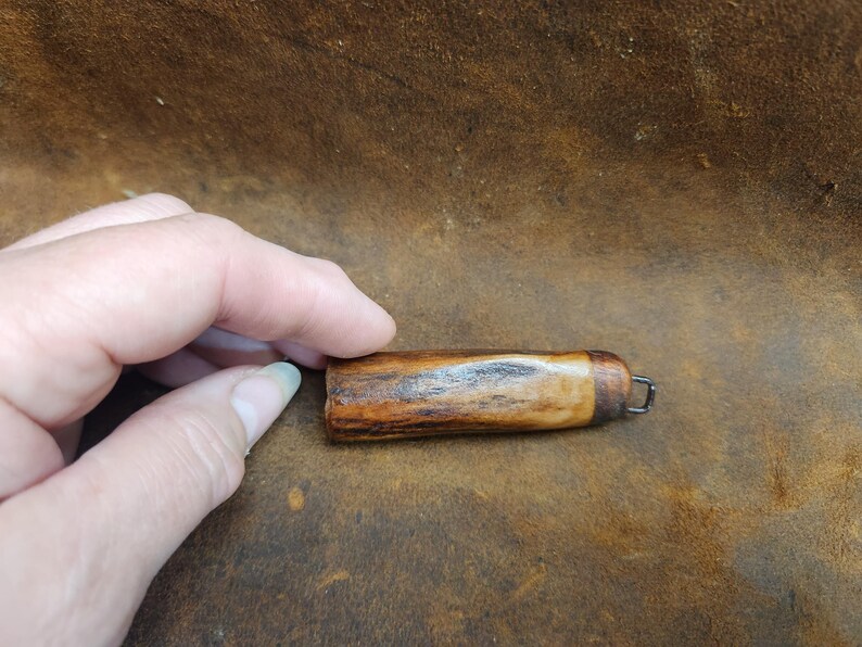 May include: A small, handcrafted wooden object with a dark brown and tan grain pattern. It has a rounded shape with a metal loop at one end. The object is held by a hand, suggesting its small size and portability.