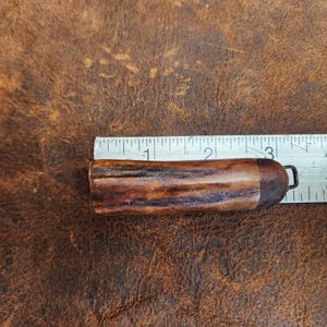 May include: A brown and tan antler object with a metal loop, next to a ruler. The antler object is approximately 3 inches long. The background is a textured brown surface.