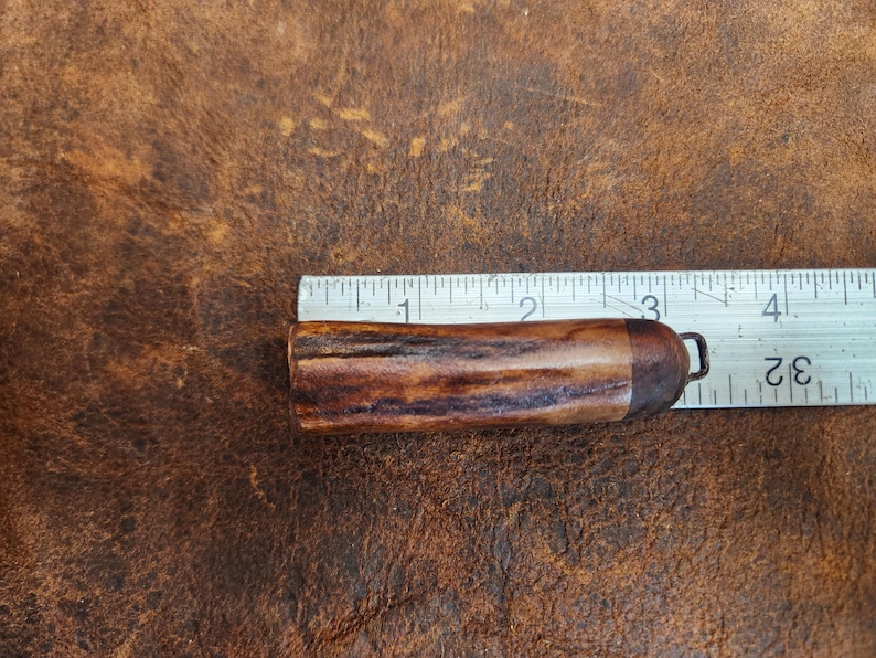 May include: A brown and tan antler object with a metal loop, next to a ruler. The antler object is approximately 3 inches long. The background is a textured brown surface.