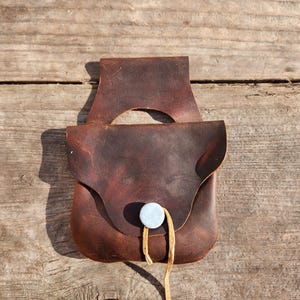 May include: A brown leather pouch with a flap closure and a loop for hanging. The pouch has a light-colored button and a leather string closure. The leather has a distressed look, and the pouch is set against a wooden background.