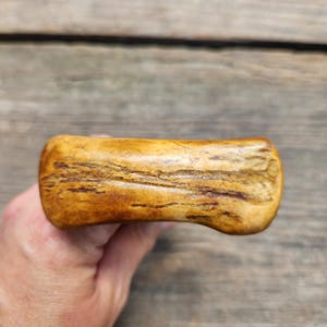 May include: A polished, rectangular piece of natural antler with a smooth, curved surface. The object features a warm, golden-brown color with darker, organic grain patterns and textured streaks running horizontally across its length.