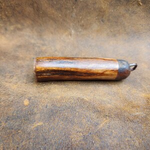 May include: A handcrafted, cylindrical bottle stopper made of wood with a dark brown and tan grain pattern. The stopper has a metal cap and a small metal loop for easy removal. The item is shown on a brown leather surface.