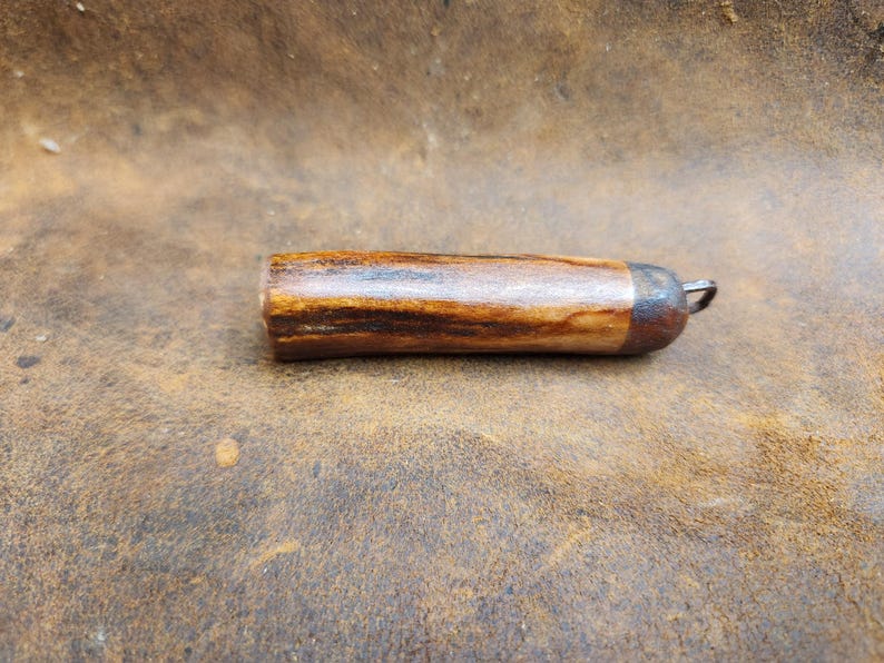 May include: A handcrafted, cylindrical bottle stopper made of wood with a dark brown and tan grain pattern. The stopper has a metal cap and a small metal loop for easy removal. The item is shown on a brown leather surface.