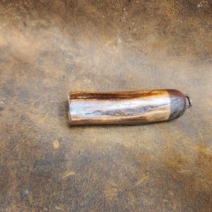 May include: A cylindrical bottle stopper made of wood and metal. The wooden portion has a light brown and dark brown pattern, with a silver-colored metal cap and a small loop for attachment. The stopper is designed to fit into a bottle.