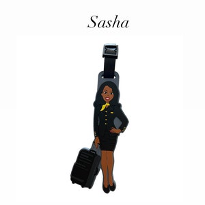 Puede incluir: Una etiqueta de equipaje con una azafata de dibujos animados con uniforme negro y bufanda amarilla, sosteniendo una maleta negra. La etiqueta tiene el nombre "Sasha" en cursiva. Está sujeta a una correa negra.