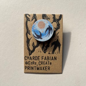 May include: A round wooden pin featuring a blue sky with a sun and two birds in flight. The pin is mounted on a brown card with the text "CHARDE FABIAN @EcRu_CREATE PRINTMAKER" in black.